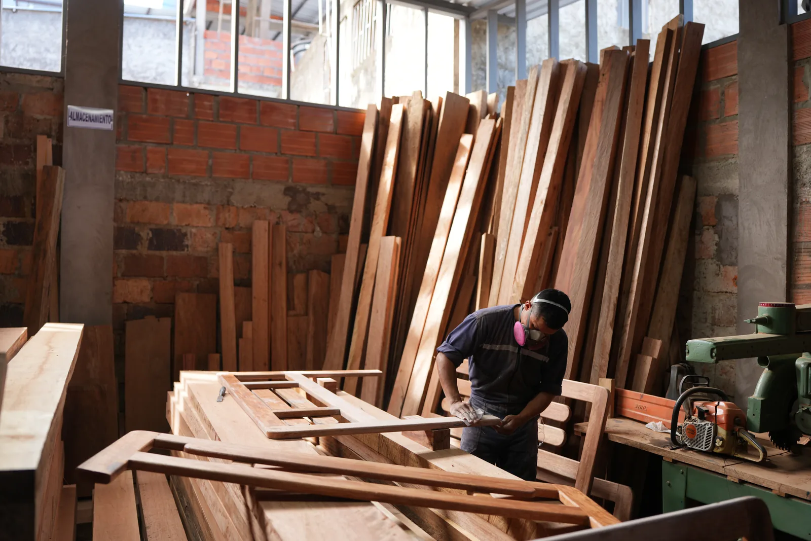 Un ebanista trabaja solo en la bodega, rodeado de tablones parados, rumas de madera y piezas de mobiliario en proceso