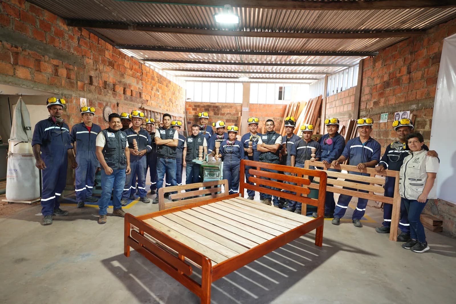 Todo el grupo de aprendices e instructores posa en el taller alrededor de una cama y una banca de madera terminadas