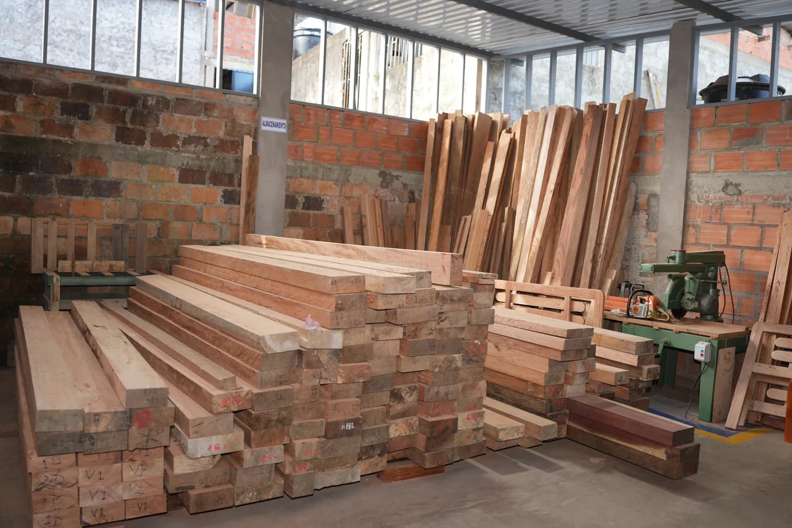 Bodega del taller con grandes rumas de madera dimensionada apilada frente a un muro de ladrillo y tablones parados contra la pared
